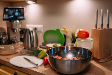 home kitchen countertop with ingredients for cooking, metal mixing bowl with minced meat, knives, vegetables, and kitchen appliances in the background