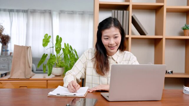 Beautiful Asian office worker, sitting working on laptop computer placed on table in front, with carefree expression, reached out hand picked up pen write down main points work in notebook.