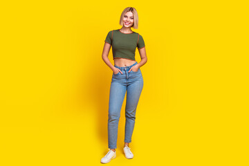 Full length photo of adorable pretty girl dressed khaki t-shirt jeans holding palms in pockets isolated on yellow color background