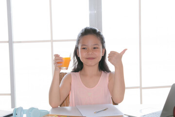 Asian Elementary school girl smiling and giving a thumbs up while holding glass of orange juice. Studying online with a laptop in a bright home environment, showing enjoyment and healthy habits.