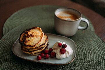 plate of homemade syrniki with whipped cream and cranberries, served with a cup of coffee on green woven placemat in cozy breakfast setting