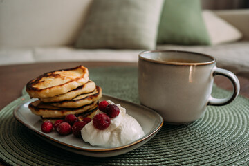 plate of homemade syrniki with whipped cream and cranberries, served with a cup of coffee on green woven placemat in cozy breakfast setting