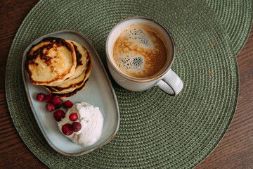 plate of homemade syrniki with whipped cream and cranberries, served with a cup of coffee on green woven placemat in cozy breakfast setting