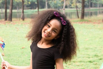 Fototapeta premium Close-up of an African-American girl with Afro curly hair style, playfully blowing on a colorful pinwheel in an outdoor park. captures joyful and curious expression, holiday recreation elementary kid