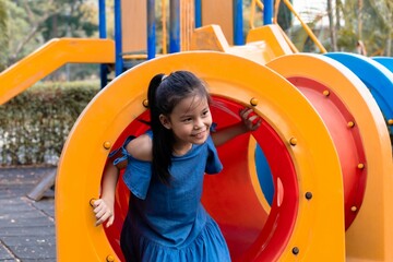 Young Asian elementary schoolgirl crawling through a colorful tunnel on a playground. weekend carefree holiday in park. bright playground equipment and focus expression capture childhood exploration