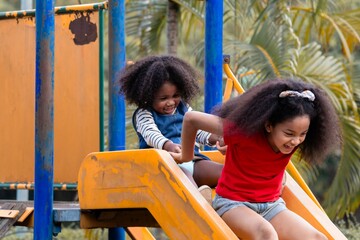 Two African American sibling girls joyful and laughing sliding down a yellow playground together,...