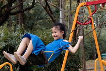 Young Asian girl enjoying a ride on a swing at the playground, smiling widely as she swings back and forth. She is wearing a blue dress and appears carefree and joyful, weekend outdoor activities