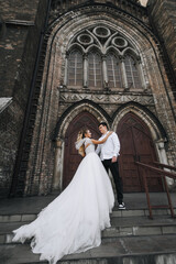 Stylish groom and beautiful blonde bride in white lace dress hugging standing against the background of old brick church. Wedding photo of happy newlyweds.