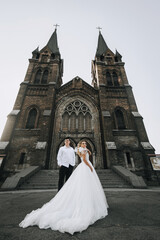 Obraz premium Stylish groom and beautiful blonde bride in white lace dress hugging standing against the background of old brick church. Wedding photo of happy newlyweds.