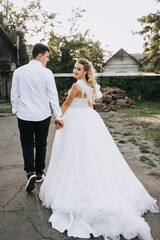 Naklejka premium Young groom and beautiful curly bride in white long dress walking holding hands outdoors against the background of logs in the village. Wedding photography, portrait of happy newlyweds.