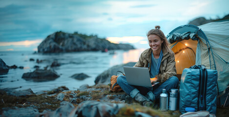 Woman camping by the ocean, using a laptop near her tent at dusk. The scene conveys a peaceful outdoor atmosphere.