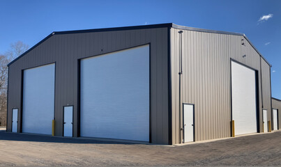 A large, brown metal building with three garage doors sits on a gravel lot with a blue sky overhead