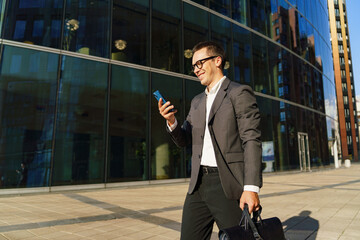 Businessman Walking With a Smartphone and Briefcase Near a Modern Glass Building on a Sunny Day