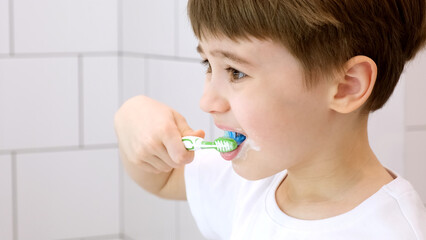 A cute little child brushes teeth. Green toothbrush. Daily morning hygiene procedures in a white bathroom. Portrait of beautiful caucasian 6 year old boy. Teeth brushing concept. Copy space. Close-up