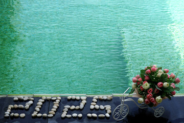 Happy New Year, 2025 seven segment numbers form written by stones on a floor tile of swimming poolside with artificial roses flower pot.