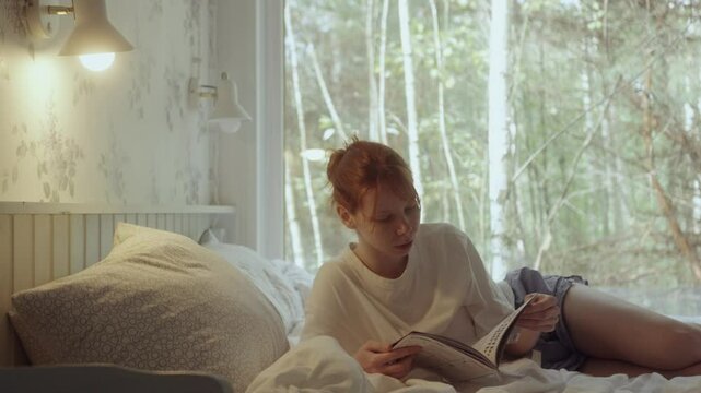 Red hair young woman in pajamas lying on her bed and reading a book, forest view