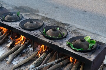 A traditional Indonesian serabi-making process, showing hands pouring batter onto a clay pan over a wood-fired stove. The smoky atmosphere and authentic tools emphasize the local street food culture.