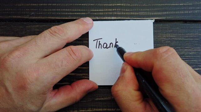 A man writes thank you and a smiley on a sticker on a dark table background