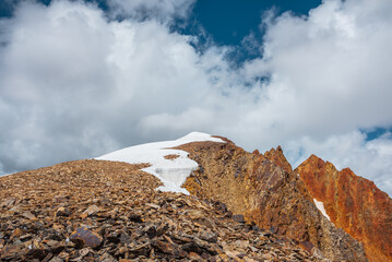 Snow-capped mountain ridge with cornice and red sharp rocks on top in sunlight. Snow cap and shiny pointy peak of gold color. Snow dome and colorful rocky peaked top. Sunny cloudy high mountains view.