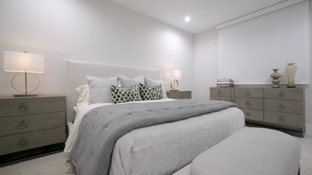 Stylish white walled bedroom interior design with multiple patterned pillows on bed and decorative table lamps on grey commodes