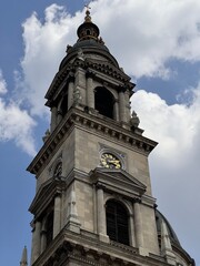A grand clock tower with ornate stonework and golden details stands tall against a cloudy sky. Statues and intricate carvings highlight the beauty of this historic structure.