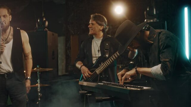 Medium shot of four Caucasian men playing musical instruments and singing at smoky studio or live concert, enjoying groove and vibe of performance
