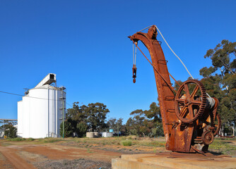 Old, rusty, historic, mechanical crane next to unused railway tracks, with grain silos in background. Clear blue sky, Western Australian Wheatbelt region. Outback.