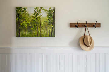 A wooden coat rack hangs on the wall, and there is an oil painting of green trees hanging above it. The white wainscoting below has a hat hanging from one hook.