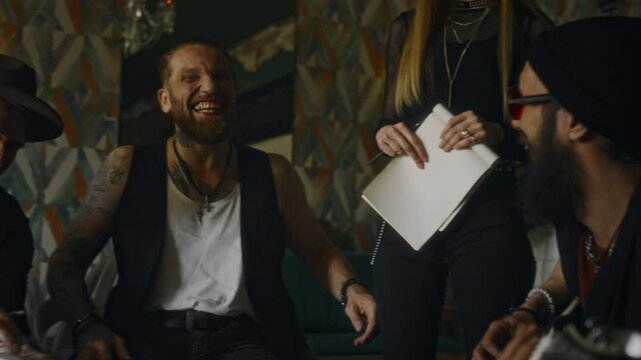 Medium slowmo shot of group of diverse musicians laughing and having fun while sitting around table in bar, playing guitar and chatting