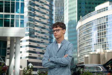 A young man stands near the Moscow City towers wearing glasses and looking into the distance