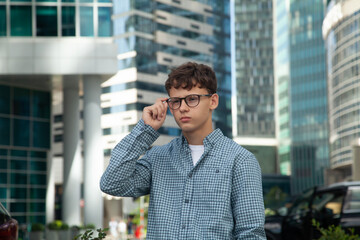 A young man stands near the Moscow City towers wearing glasses and looking into the distance