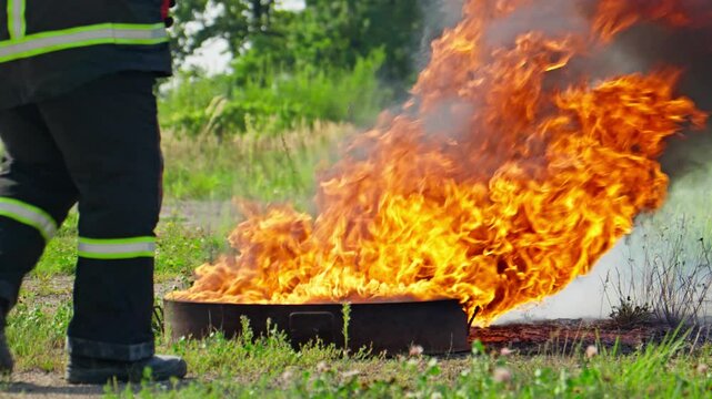 Firefighter battles roaring flame using fire extinguisher to combat spreading flames in open field surrounded by greenery. Firefighter demonstrates proper technique and safety precautions. Slow motion