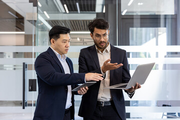 Two businessmen engaged in discussion holding laptop and tablet in modern office. Displaying teamwork, communication in professional business environment. Emphasis on collaboration, technology