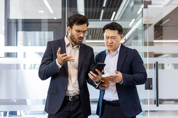 Two businessmen in modern office setting intensely discuss news displayed on smartphone. Colleagues...