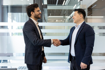 Two businessmen exchange a confident handshake in modern office environment, showcasing professional interaction and teamwork. They stand facing each other, wearing formal attire