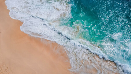 A serene aerial view of waves gently rolling onto a pristine sandy beach. The turquoise ocean meets the soft golden sand, creating a stunning contrast between land and sea in this tranquil coastal
