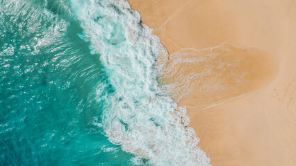An aerial shot capturing turquoise ocean waves meeting golden sands at Kelingking Beach, Nusa Penida, Indonesia. Subtle footprints can be seen along the pristine shoreline.
