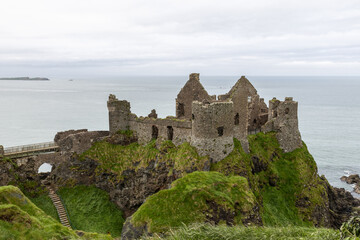 Fototapeta premium Dunluce Castle sits on a unique rocky cliff overlooking the ocean, with steep green slopes and rugged terrain. This historic landmark is framed by the sea and cloudy sky