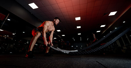 Fototapeta premium Man exercising with battle ropes in a gym at night. A shirtless man engages in an intense workout using battle ropes inside a dimly lit gym during evening hours.