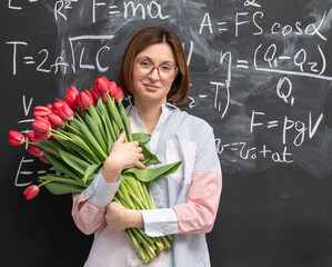 A young attractive teacher stands with a bouquet of flowers in the classroom. Teachers day and graduation concept