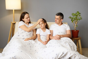 Unwell little girl lying in bed with her parents child suffering high fever while mom checking her health by touching forehead family have illness posing in cozy home