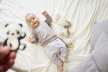 Obraz premium Child’s innocence. Relaxed childhood. Caucasian baby lying on bed in bedroom resting happily while unrecognizable mother showing tiny toy to infant son