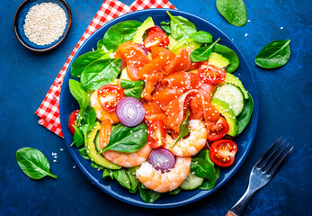 Salmon salad for ketogenic diet with shrimp, avocado, spinach, cucumber, tomato, cashew nuts, sesame. Low-carbohydrate lunch rich in healthy fats. Blue table background, top view