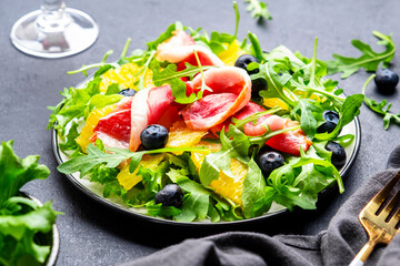 Foodie food salad with raw smoked duck and juicy orange, blueberry and arugula on black background, top view