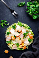 Caesar salad with grilled chicken slices, iceberg lettuce, croutons, parmesan cheese and classic dressing. Black background, top view