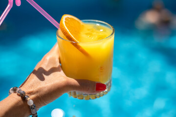 A glass of orange lemonade with a slice of orange and ice in hand against the background of blue pool water
