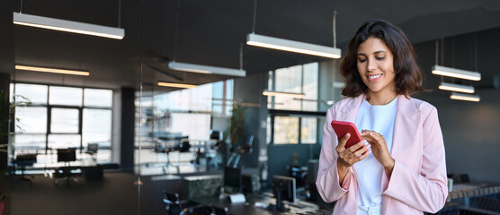 European business woman manager using cell phone mobile application standing indoors in office. Latin Hispanic young female businesswoman entrepreneur focused working on smartphone. Banner, copy space