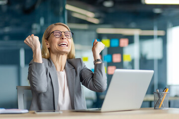 Joyful businesswoman in office celebrating achievement with clenched fists. Displays excitement and success. Sitting at desk with laptop, this professional embodies career accomplishment