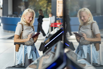 The female traveler checks her phone while interacting with an railway station kiosk, preparing for...