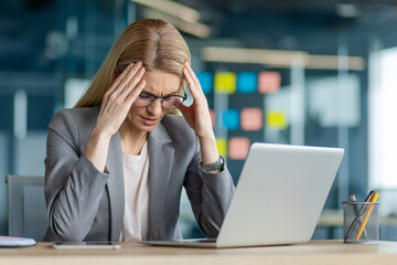 Businesswoman in gray suit at desk feeling stressed while working on laptop. Struggles with work pressure and challenges in modern office environment, expressing frustration and concern.